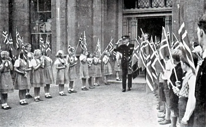 Schoolchildren holding flags greet a uniformed man outside a building, a historic scene of schools worldwide during World War II.