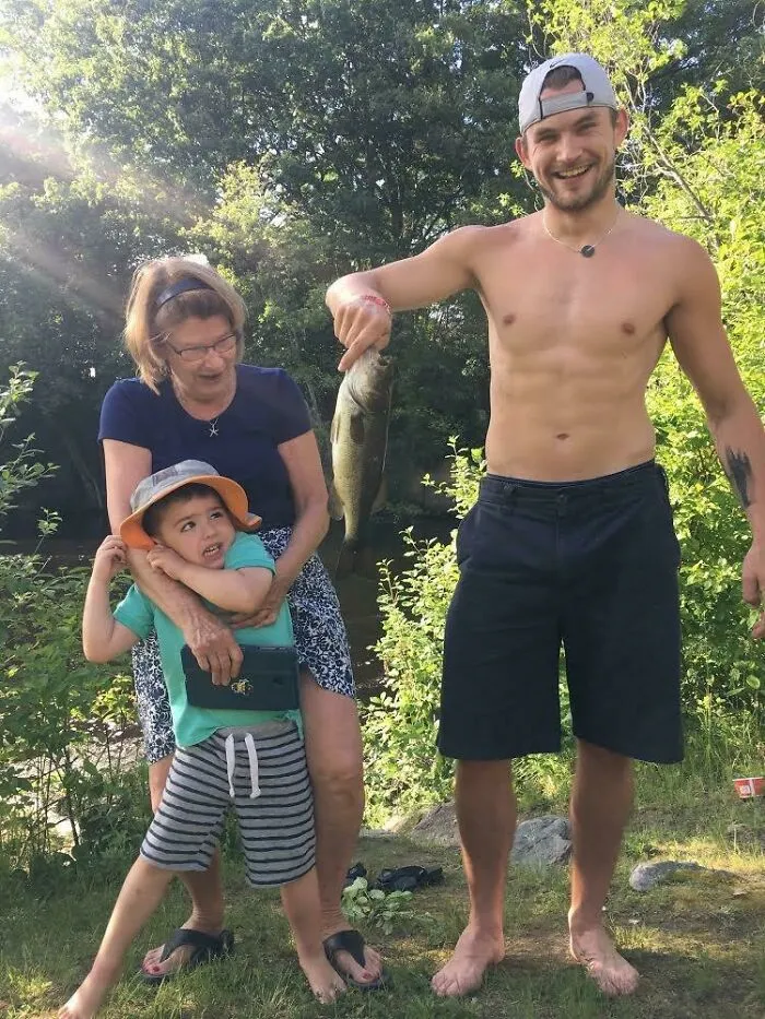 Hombre sonriente mostrando pez pescado en vacaciones de verano con niño y mujer mayor en un entorno natural divertido.