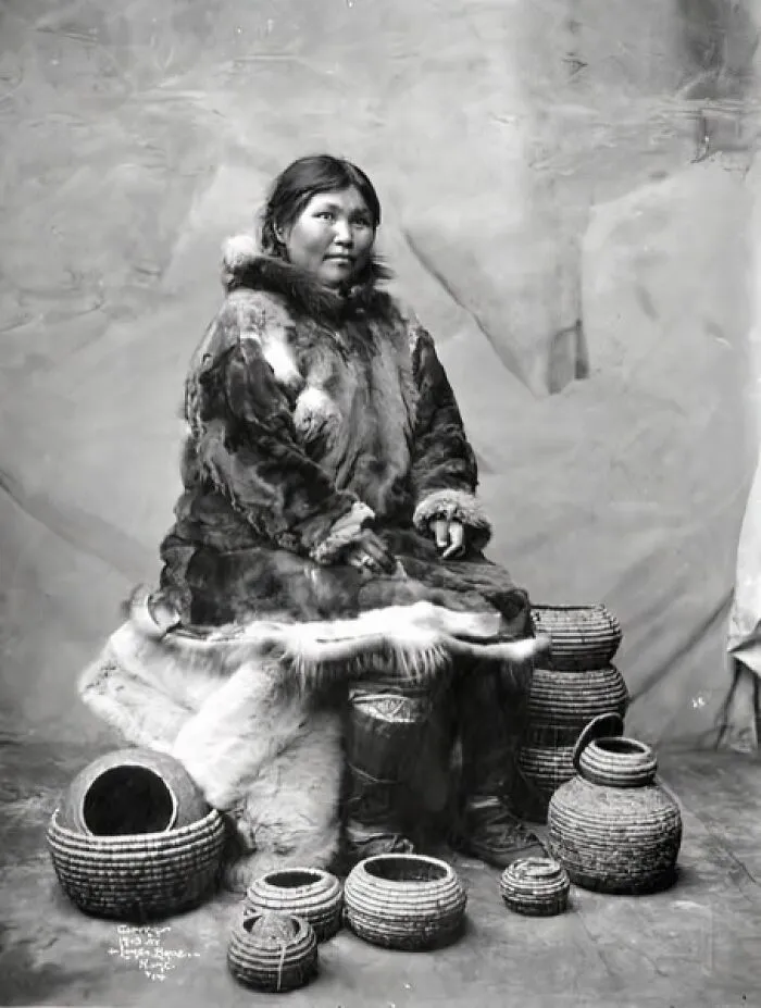 Studio Portrait Of Eskimo Woman Dressed In Moose Fur Parka With Seal Bladder Boots Seated With Several Coil Baskets, Nome, 1903