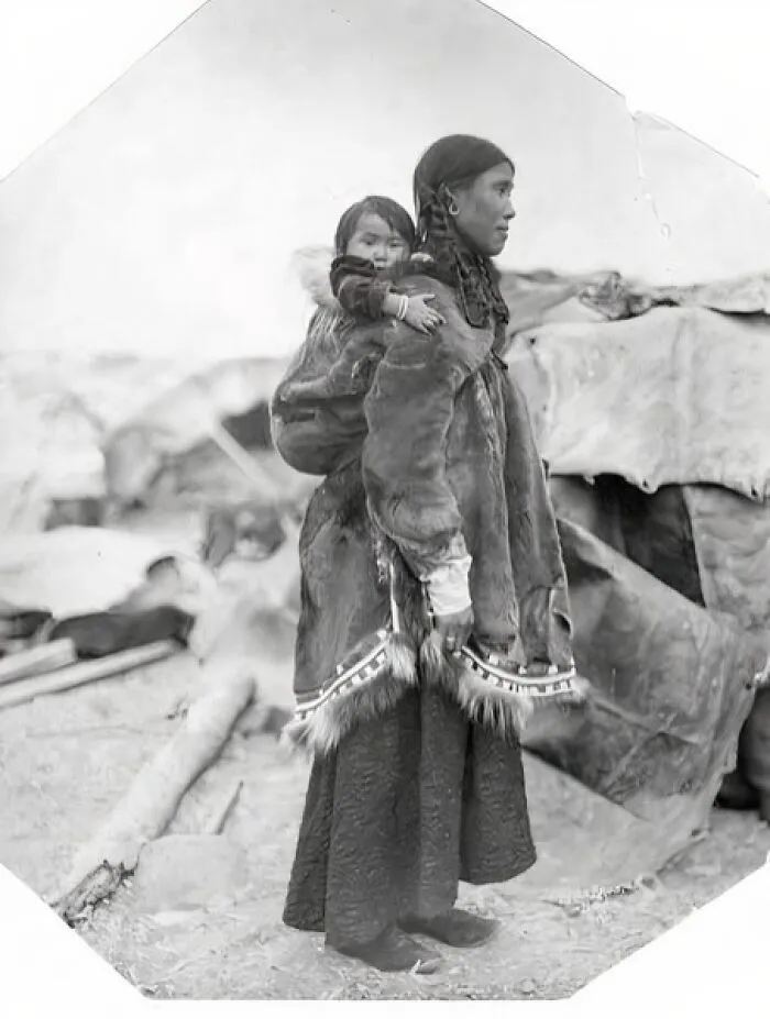 Inuit Mother Wearing Long Fabric Skirt And Fur Parka, Standing With Baby On Her Back Near Encampment, Nome, Alaska, Between 1905 And 1910