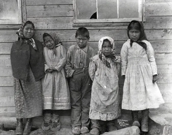 Historic black and white photo of Inuit children standing in front of a wooden building, capturing Inuit spirit and resilience.