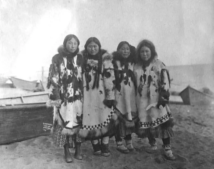 Historic photo of four Inuit women wearing traditional fur clothing standing together near boats on a beach, showcasing Inuit spirit.