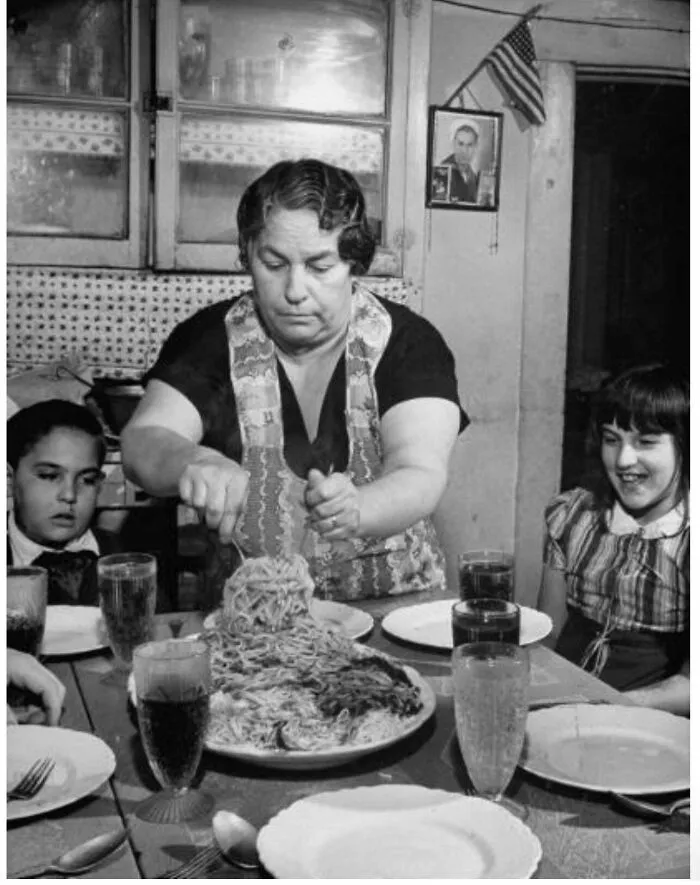 Woman serving a large plate of spaghetti to children at a dining table, a candid glimpse into the past.