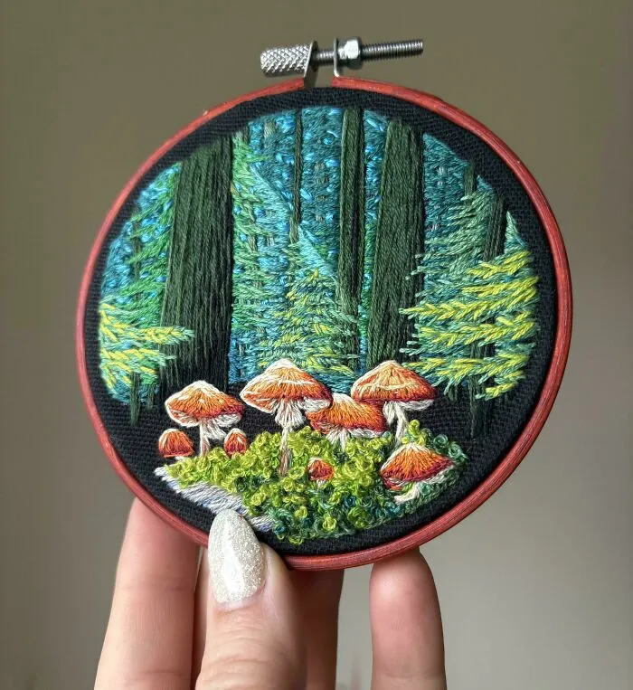 Hand holding embroidery hoop with detailed forest scene featuring mushrooms and trees, showcasing embroidery skills.