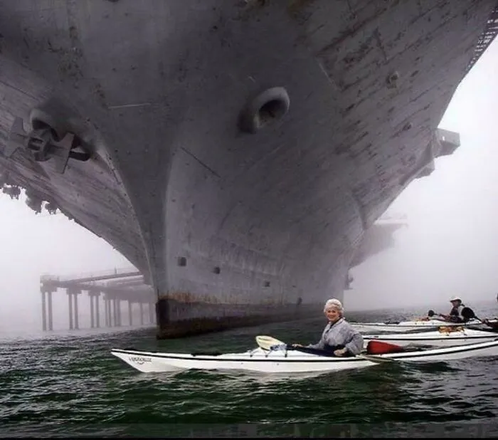 A small kayak with a person paddling near the enormous bow of an extremely large ship emerging from heavy fog, an example of how large things can be eerie.
