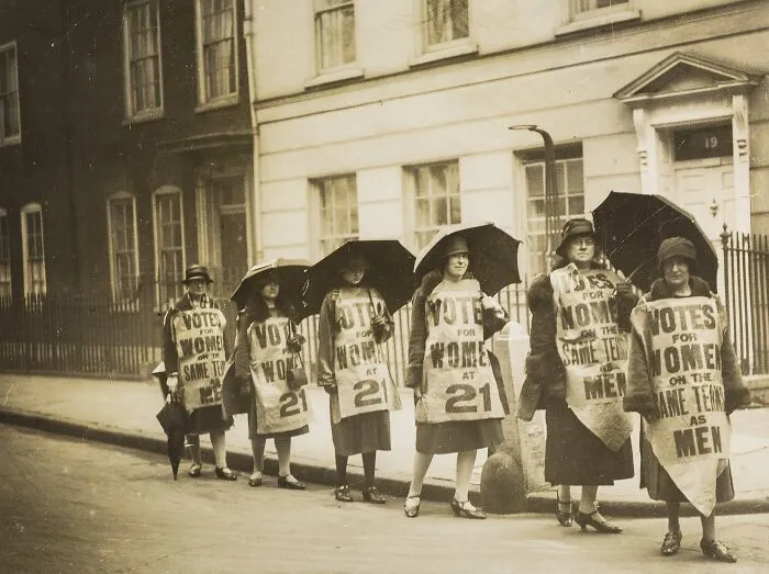 Six Women In A Line In A Street, Five Carrying Umbrellas, Wearing Full Length Tabards With Slogans 'Votes For Women On The Same Terms As Men' And 'Votes For Women At 21', 1927