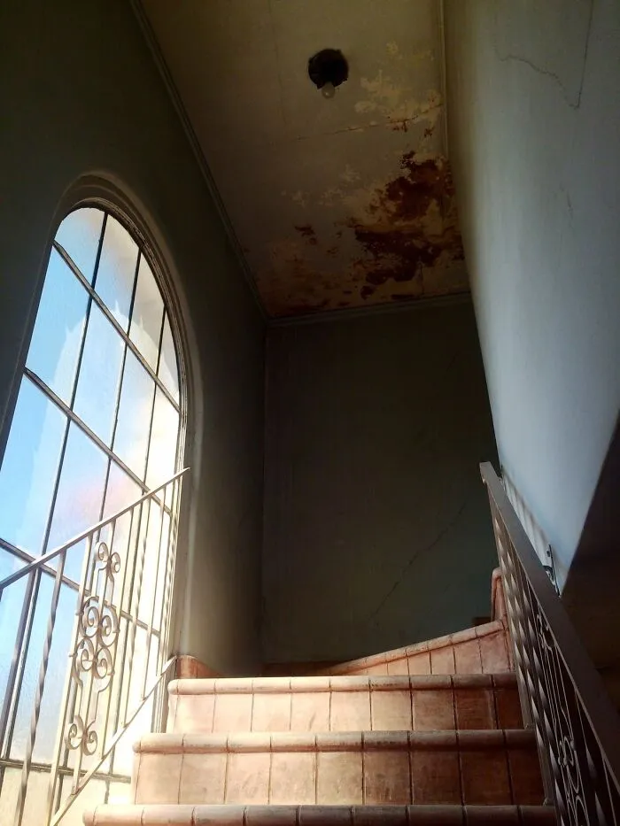 Abandoned places interior showing worn staircase, cracked walls, rusty ceiling, and a large arched window with natural light.