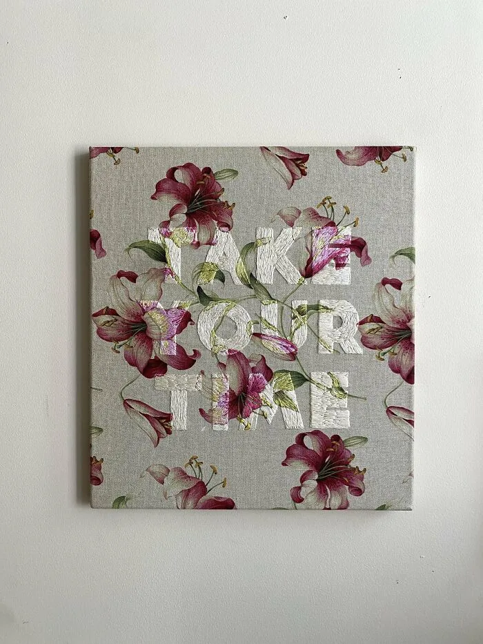Floral fabric canvas embroidered with white and colored thread forming the words take our time, showcasing embroidery skills.