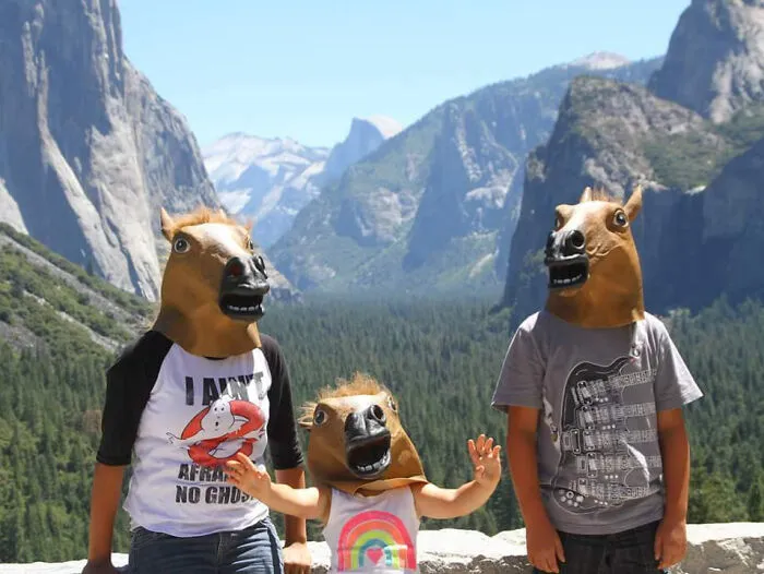 Tres personas con máscaras de caballo en vacaciones de verano divertidas en un parque natural con montañas y bosques de fondo.