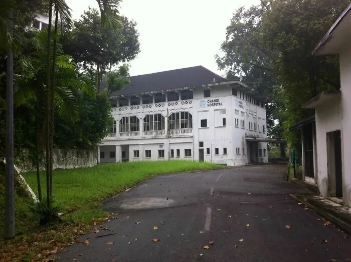 Abandoned Changi Hospital building surrounded by overgrown grass and trees on a deserted road.