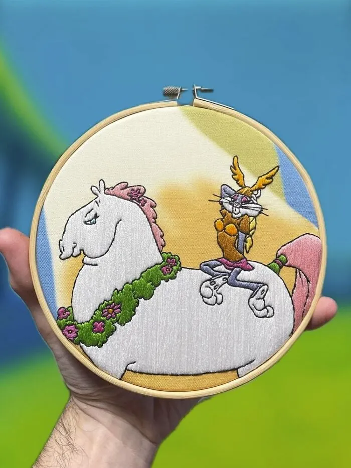 Hand holding an embroidery hoop with a colorful stitched design of a white horse and a cartoon character showing embroidery skills.