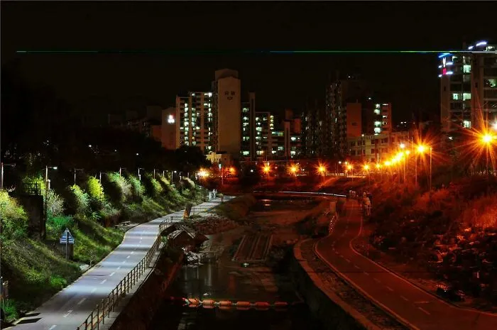 Camino iluminado junto al río en ciudad nocturna, mostrando comparaciones y diversidad inesperada de la vida urbana.