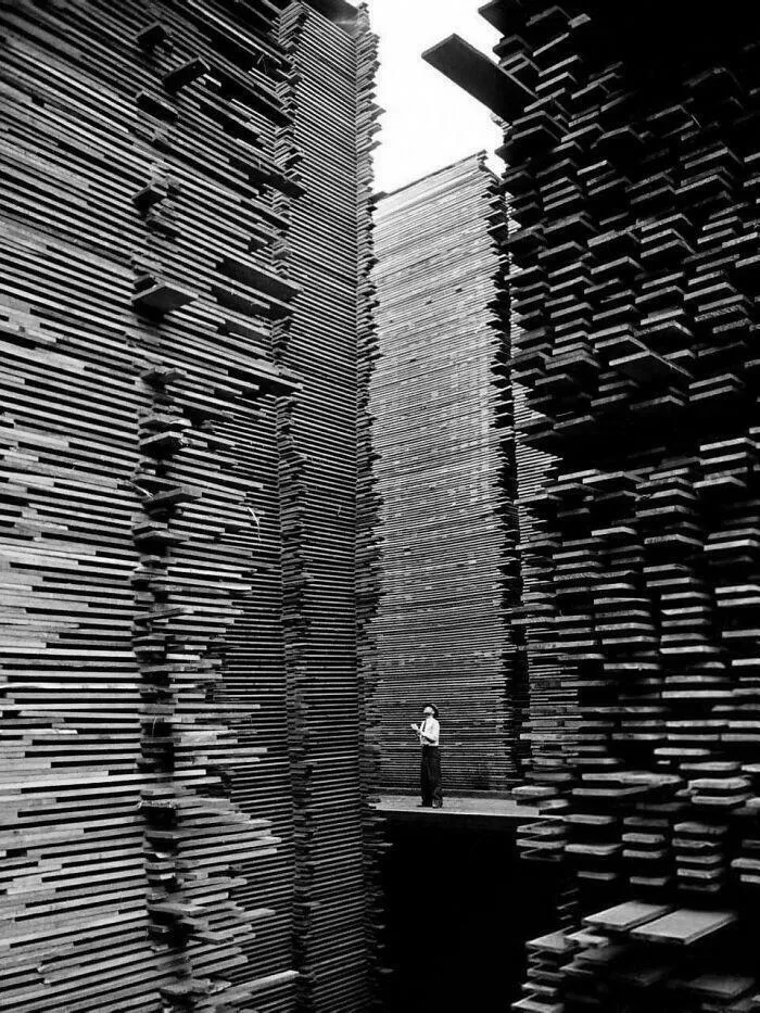 A person standing between two towering stacks of wood planks, creating an eerie sense of extremely large things.