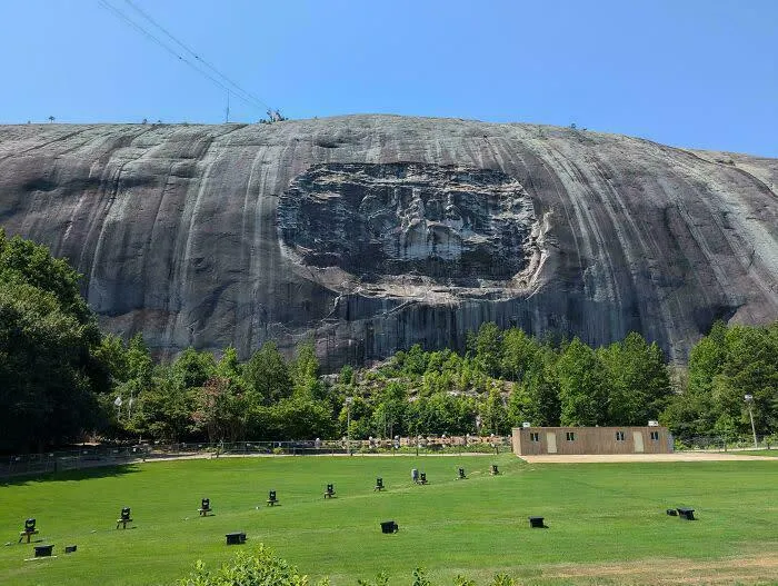 A carved mountain showing extremely large things, green grass, and trees under a clear blue sky, eerie.