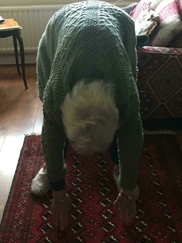 Elderly person in a green sweater touching toes on a patterned rug, a moderately interesting moment shared with the world.