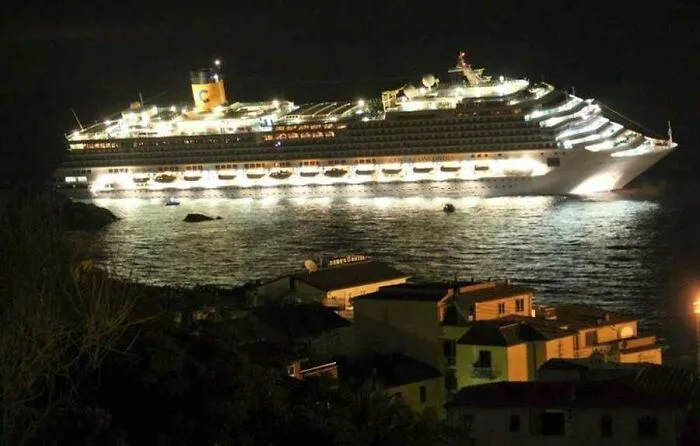 Extremely large things: A giant cruise ship, brightly lit at night, looms over small coastal houses, looking scary and eerie.