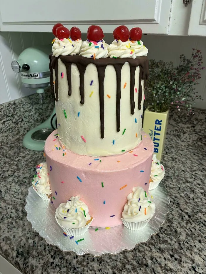 Two-tiered cake with chocolate drip, whipped cream, cherries, and colorful sprinkles, surrounded by mini cupcakes, baked goods display.