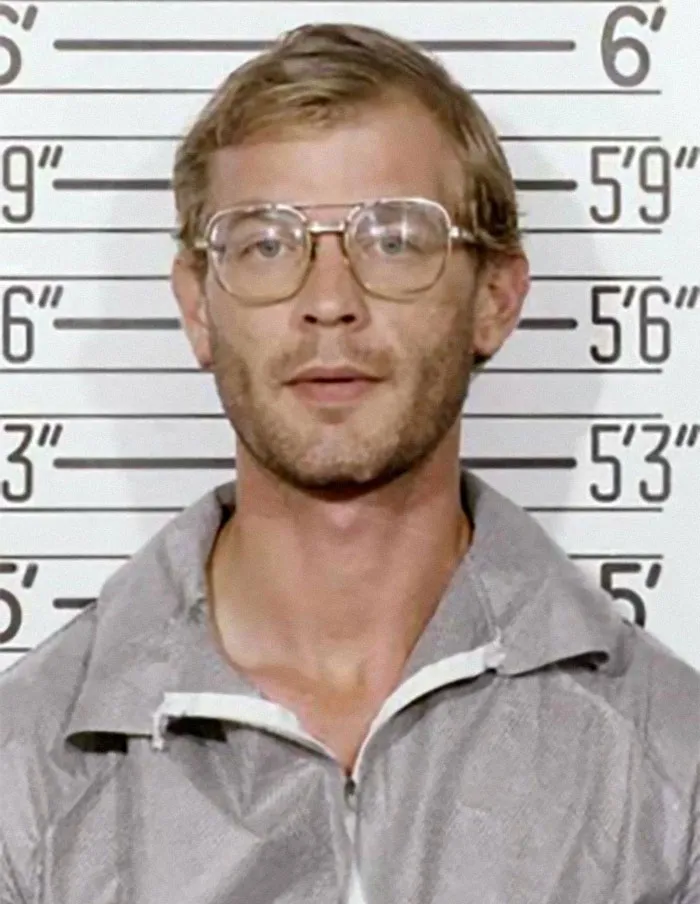 Mugshot photo of a man with glasses and casual jacket in front of a height chart, part of family flexes collection.