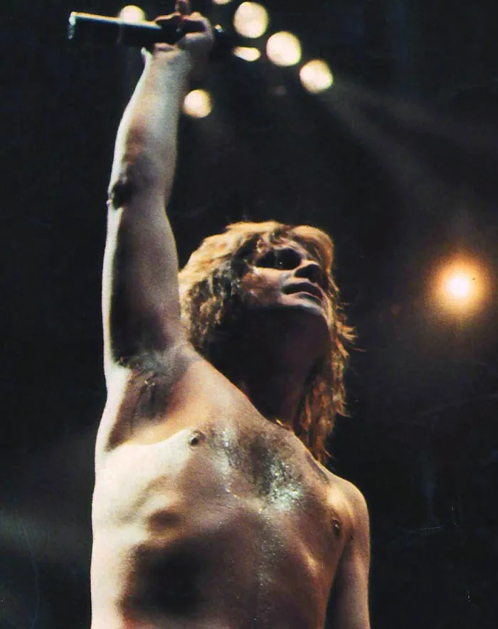 Shirtless male rock performer on stage holding microphone up, showcasing insane family flexes with intense lighting.