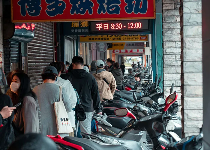 People waiting in line at a busy street market with motorcycles parked along the sidewalk, illustrating everyday life scenes.