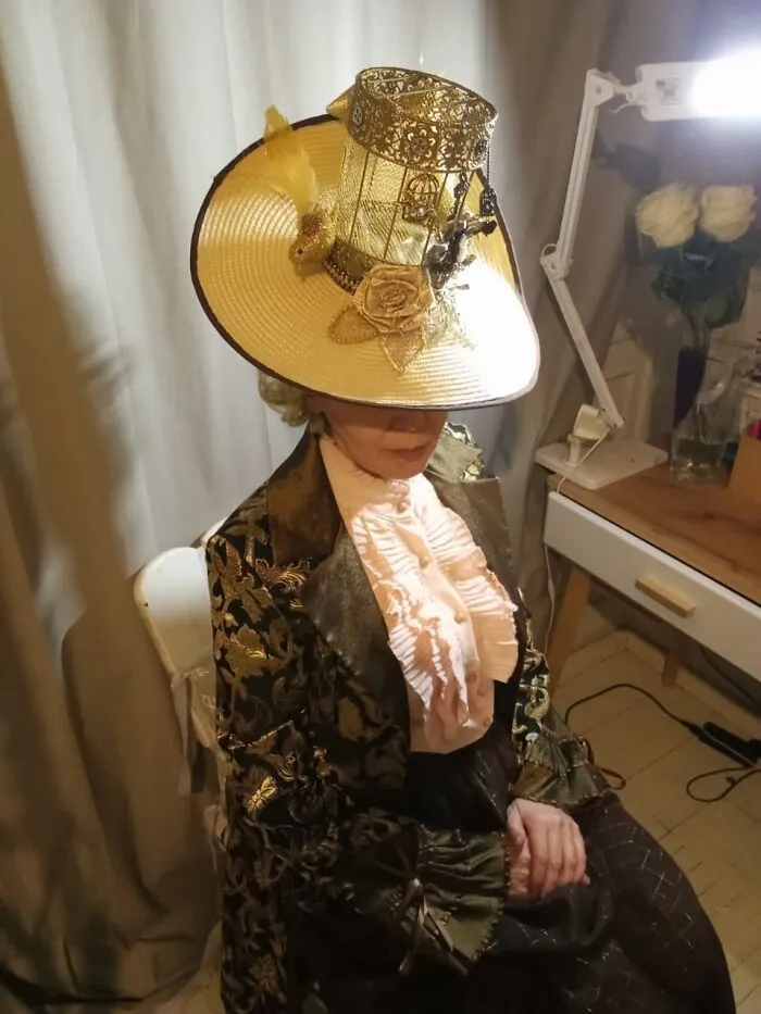 Woman in an incredible cosplay wearing a decorative hat and vintage-style clothing, sitting indoors by a desk with flowers.