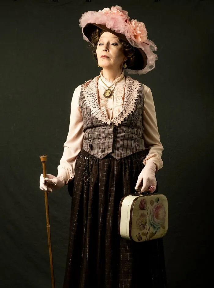 Woman in incredible cosplay wearing vintage outfit with a large pink hat, holding a cane and floral handbag against dark background