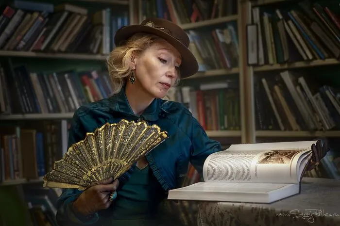 Woman in vintage cosplay outfit with a hat and fan, reading a large book in a library setting, showcasing incredible cosplays.