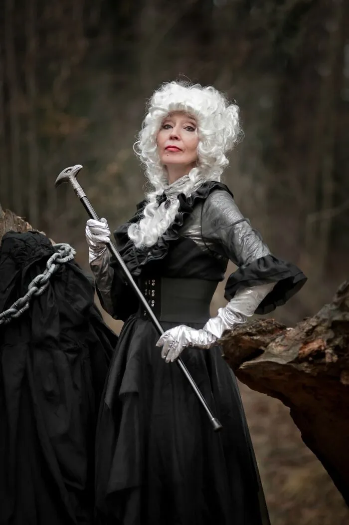 Woman in an incredible cosplay wearing a white wig and black Victorian-style dress holding a cane outdoors in a forest setting