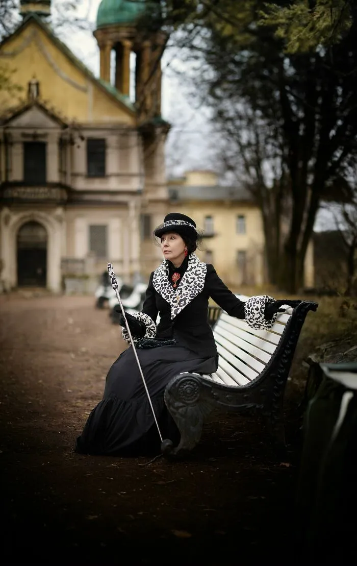 Woman in incredible cosplay with leopard print accents sitting on a park bench holding a cane in front of old building.
