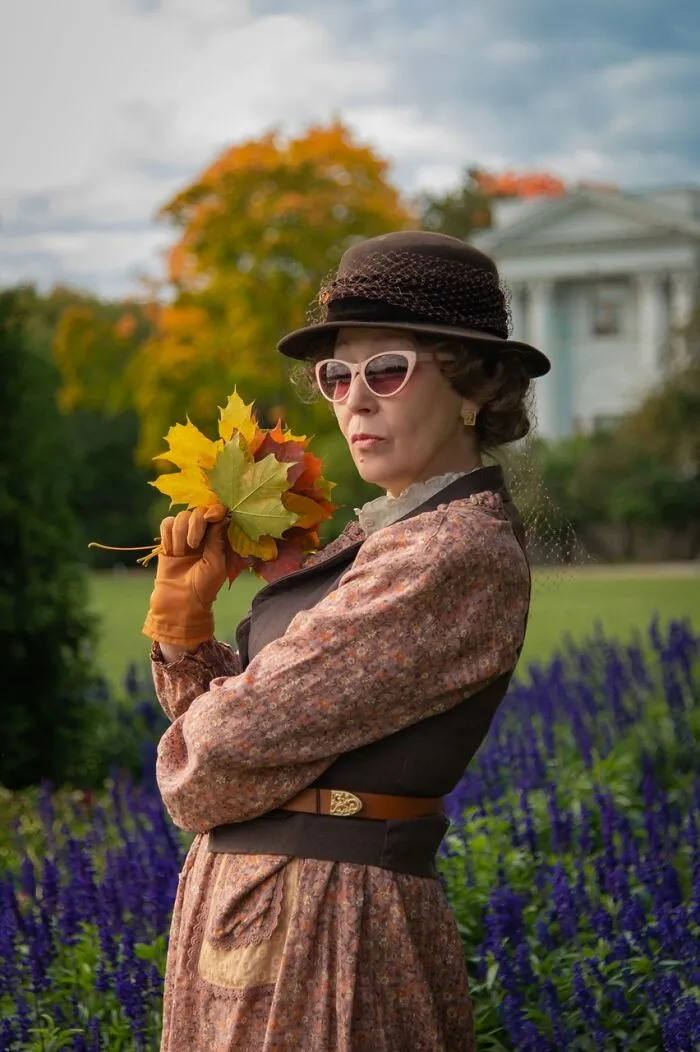Woman in vintage cosplay outfit holding autumn leaves, wearing hat and sunglasses in a garden with flowers and trees.