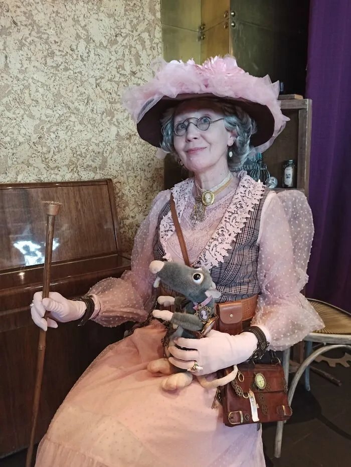 Woman in an incredible cosplay wearing a vintage pink dress and hat, holding a puppet and a walking cane indoors.