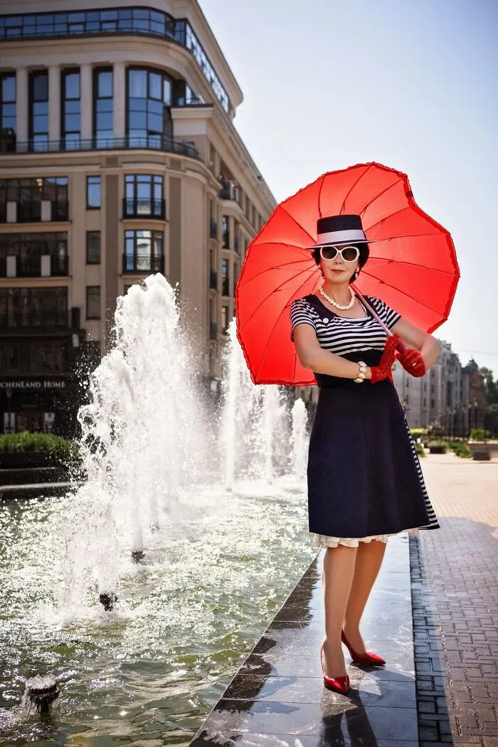 Woman in vintage cosplay outfit with red umbrella and gloves posing near fountain in urban setting.