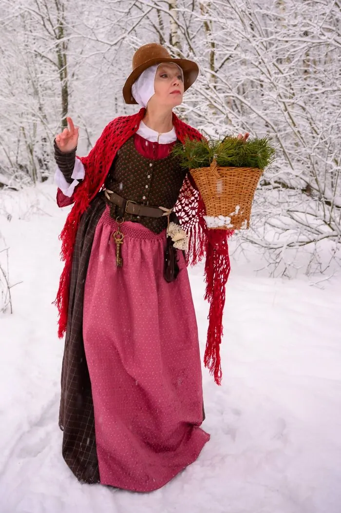 Woman in incredible cosplay wearing vintage dress and red shawl, carrying basket in snowy forest setting.