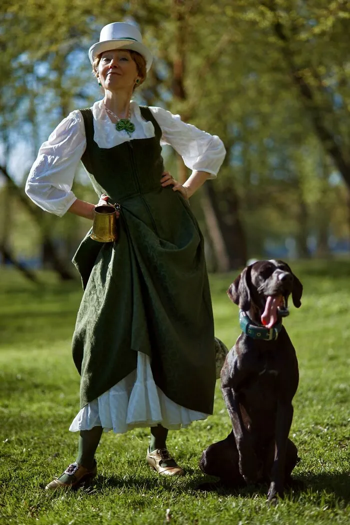 Woman in an incredible cosplay outfit with a green dress and white hat standing outside with a large black dog.