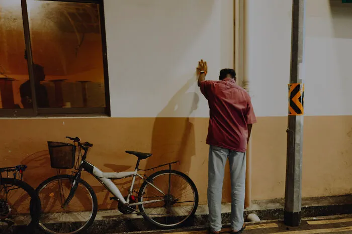Man wearing a red shirt leaning against a wall next to a bicycle in a poetic, cinematic image by a photographer.