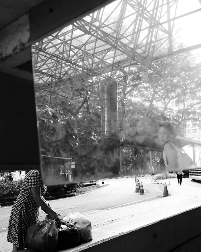 Black and white cinematic image showing a woman handling bags and a reflective surface capturing an urban scene and a pedestrian.