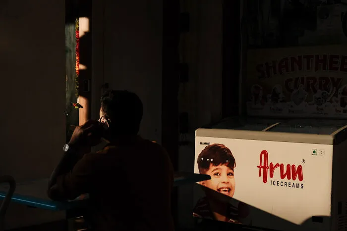 Man sitting in dim light beside an ice cream freezer, captured in a poetic, cinematic style by the photographer.