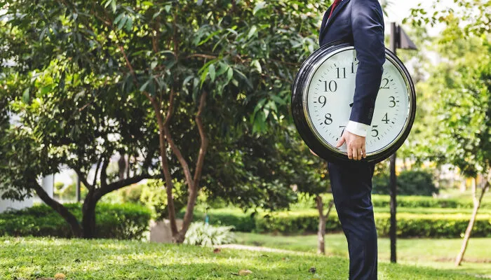 Man in a suit holding a large clock outdoors, illustrating concept time facts in a green park setting.