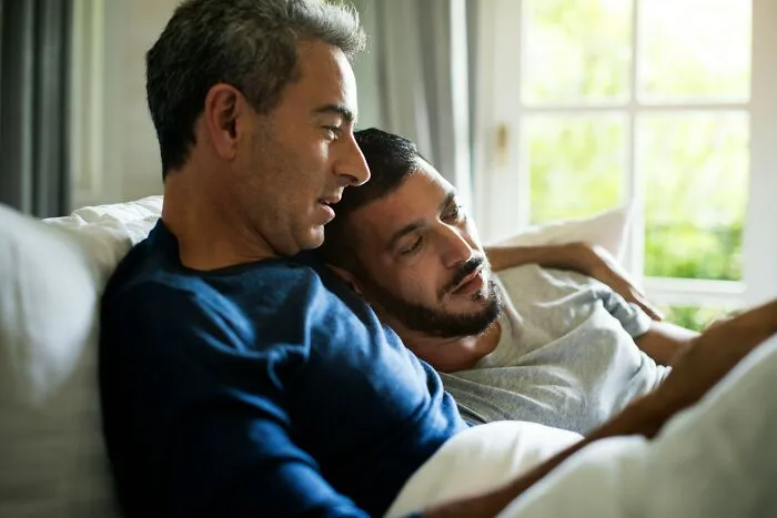 Two men sitting closely on a couch, sharing a quiet moment while discovering hidden family secrets.