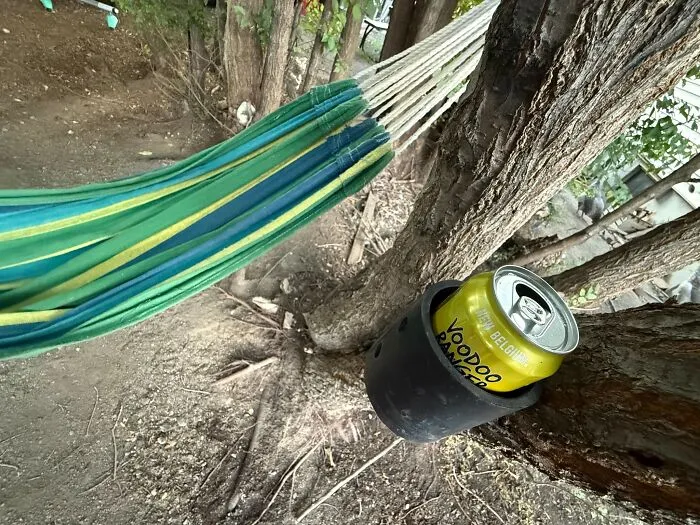 Green and blue hammock tied between trees with a cold drink can resting in a holder on a tree outdoors, appealing to men.