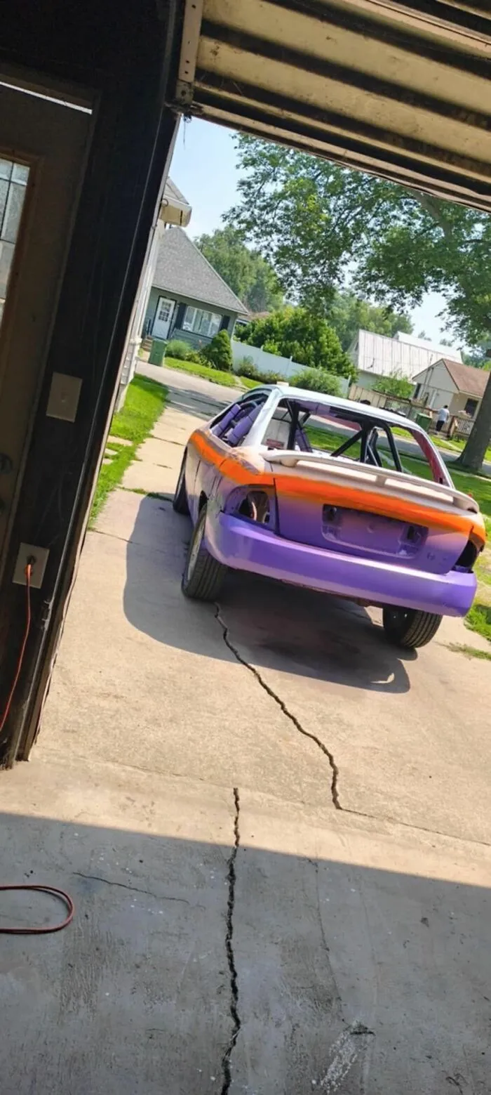Purple and orange car body in driveway, viewed from garage with cracked concrete, surrounded by suburban houses and trees.
