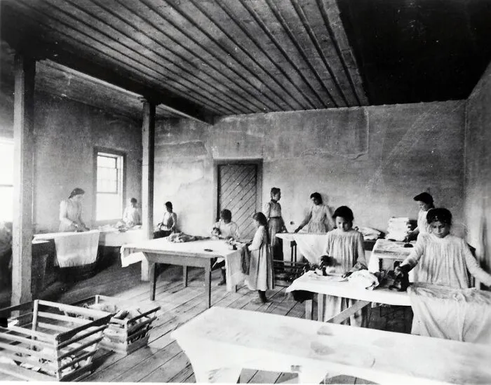 Native American boarding school students in the 1910s engaged in chores inside a sparse classroom setting.