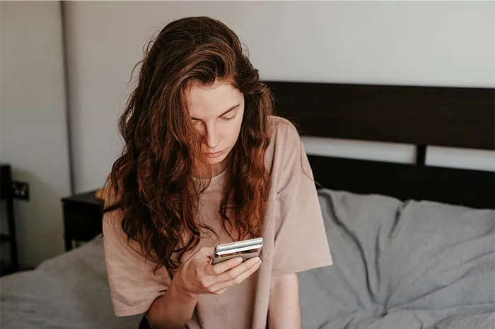 Young woman with long hair sitting on a bed, looking at her phone, reflecting on the Boston Marathon experience.