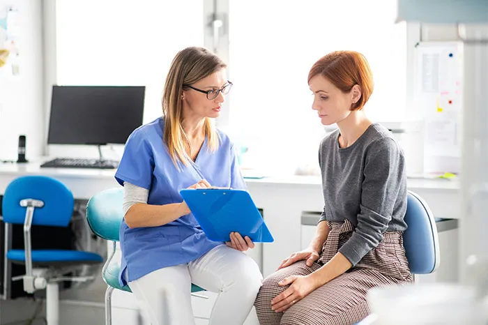 A nurse in blue scrubs discussing health details with a female patient in a bright medical office setting.