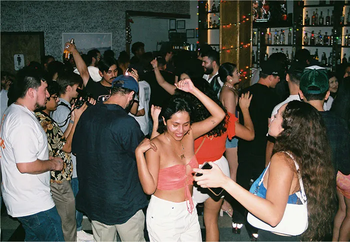 Crowd of people enjoying a lively gathering, with many dancing and socializing at a busy indoor event near a bar.
