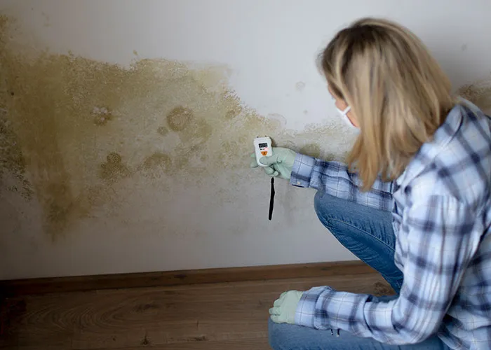 Woman wearing mask and gloves using a device to check mold on a wall, highlighting risks of patients being too young to be sick.