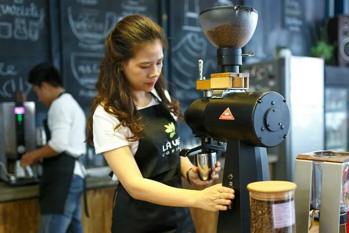 Young female barista using coffee grinder in cafe with coworker in background, illustrating stories of coworkers who are morons
