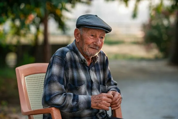 Elderly man wearing a flat cap and plaid shirt sitting outdoors reflecting on hidden family secrets discovered after a loved one passed.