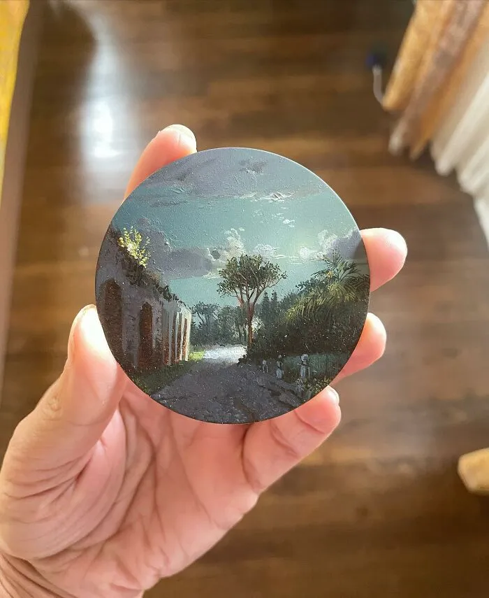 Hand holding a detailed painting by Dina Brodsky depicting a natural landscape with trees and a cloudy sky.
