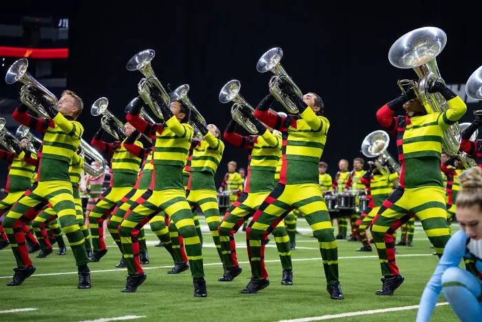 Marching band members in bright yellow and green striped uniforms performing with brass instruments on a football field.